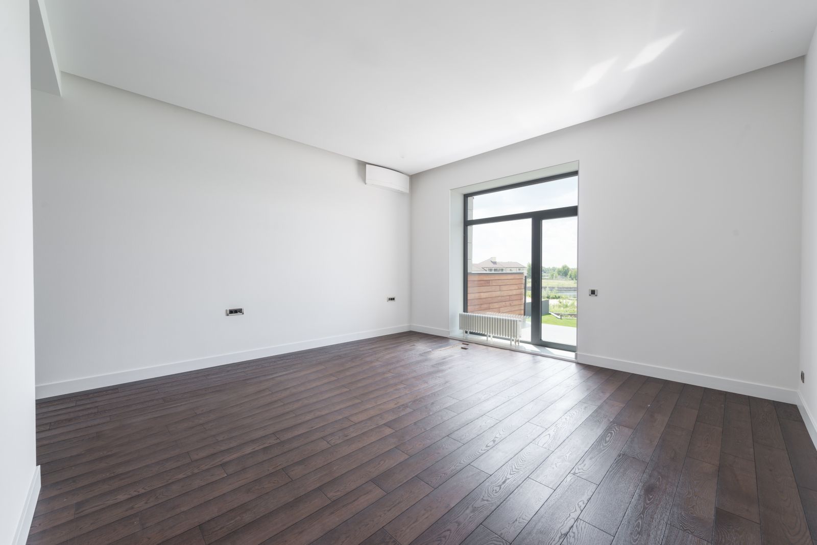 Salón vacío de una vivienda recién terminada, con suelo de madera, paredes blancas, enchufes a la vista y un gran ventanal que da a la terraza