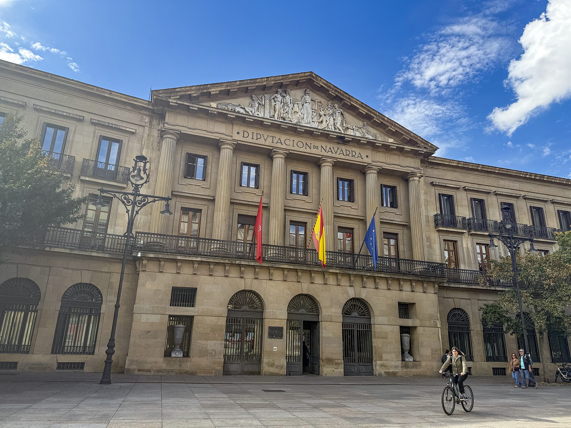 Fachada del Palacio de Navarra en Pamplona, sede del Gobierno foral, con las banderas en el balcón y una joven en bicicleta en la plaza