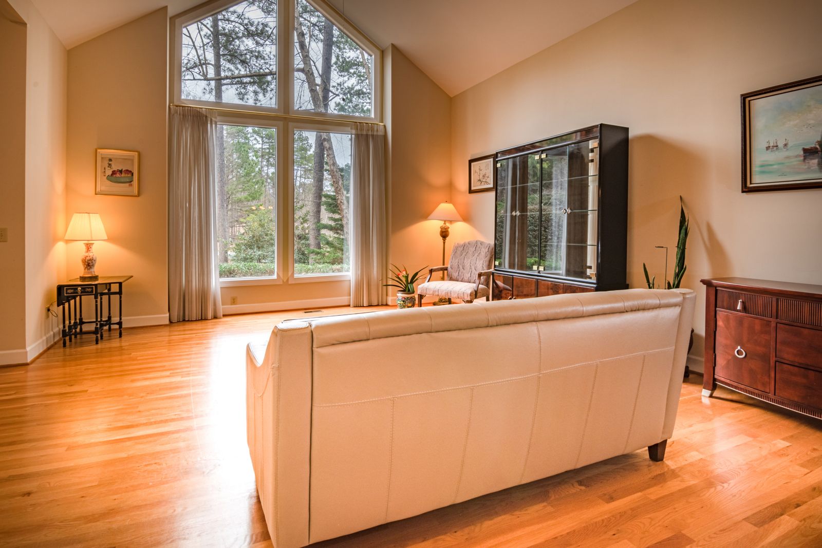 Salón luminoso de una vivienda con suelo de madera, sofá claro y un gran ventanal de doble altura que da a un jardín con árboles